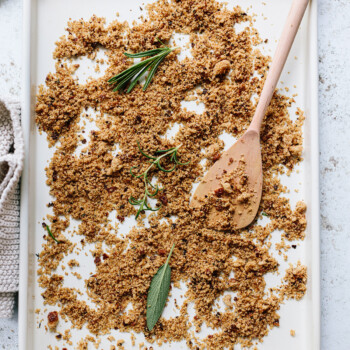 A baking sheet scattered with golden-brown homemade breadcrumbs, a wooden spoon, and sprigs of fresh herbs like rosemary and sage on a white surface.
