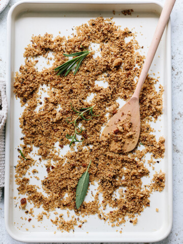 A baking sheet scattered with golden-brown homemade breadcrumbs, a wooden spoon, and sprigs of fresh herbs like rosemary and sage on a white surface.