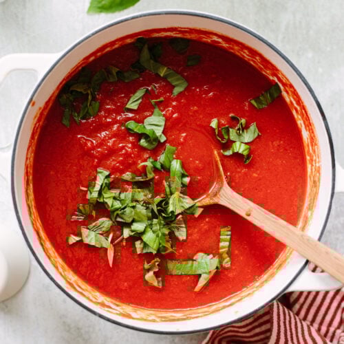 Crushed tomato marinara sauce and a wooden spoon in a white Dutch oven, topped with fresh basil ribbons.