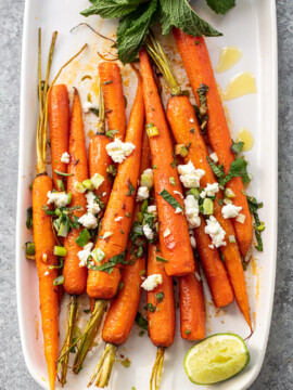 Roasted carrots on a platter, with lime wedge, fresh herbs and crumbled cheese scattered over.