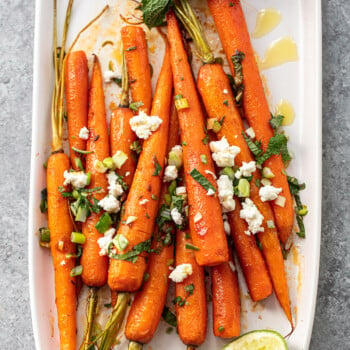 Roasted carrots on a platter, with lime wedge, fresh herbs and crumbled cheese scattered over.