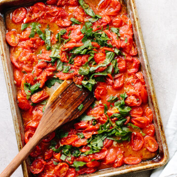 Roasted tomatoes on a sheet pan topped with fresh basil, crushed with a wood spoon.