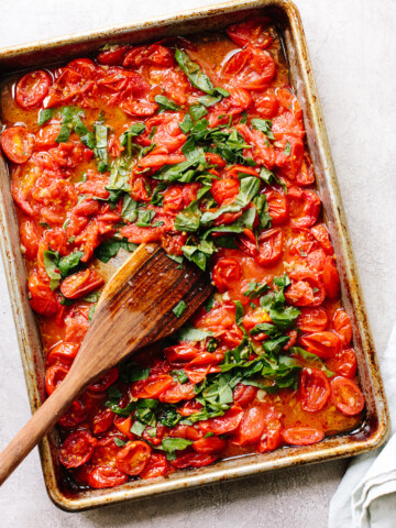 Roasted tomatoes on a sheet pan topped with fresh basil, crushed with a wood spoon.