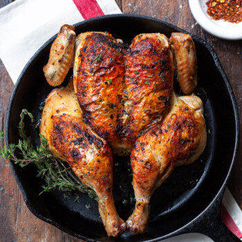 A roasted butterfiled whole chicken in a cast iron skillet, set on top of a linen napkin on a worn wood surface.