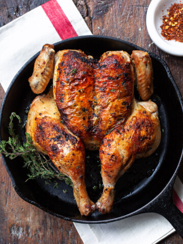 A roasted butterfiled whole chicken in a cast iron skillet, set on top of a linen napkin on a worn wood surface.