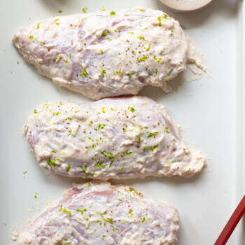 Three boneless, skinless chicken breasts coated with a thick yogurt marinade and fresh lime zest, arranged on a baking sheet.