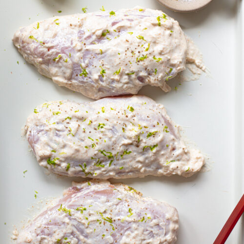 Three boneless, skinless chicken breasts coated with a thick yogurt marinade and fresh lime zest, arranged on a baking sheet.