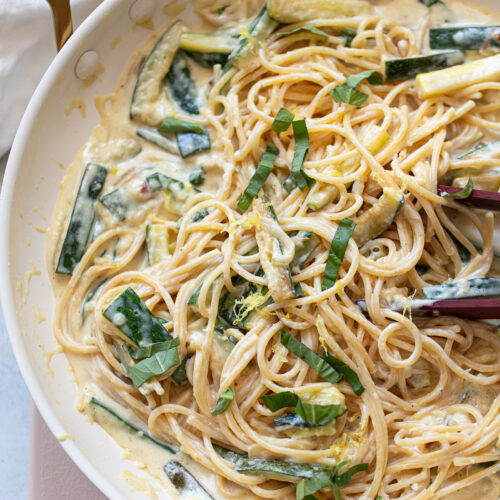 Cooked spaghetti in a skillet, sauced with cream, zucchini, lemon and basil, with a pair of tongs to the side of the pan.