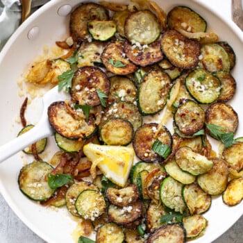 Sautéed zucchini slices with golden brown edges, garnished with fresh basil leaves, grated cheese, and a lemon wedge—an option for vegetable side dishes, served in a white skillet with a white spoon.