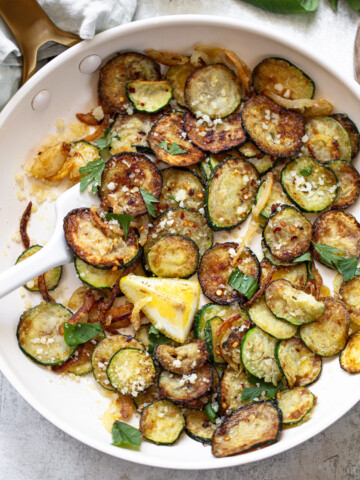Sautéed zucchini slices with golden brown edges, garnished with fresh basil leaves, grated cheese, and a lemon wedge—an option for vegetable side dishes, served in a white skillet with a white spoon.