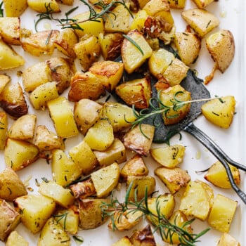 Roasted golden potatoes, cut into cubes, rest on a baking sheet with fresh rosemary sprigs. A metal spatula lifts some of the crispy roasted potatoes, ready to serve.