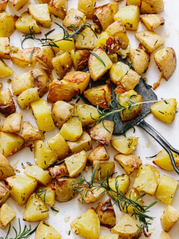 Roasted golden potatoes, cut into cubes, rest on a baking sheet with fresh rosemary sprigs. A metal spatula lifts some of the crispy roasted potatoes, ready to serve.