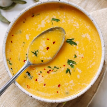 A bowl of creamy butternut squash soup garnished with herbs, Parmesan cheese and red pepper flakes, with a spoon resting on top. The bowl sits on a beige napkin.
