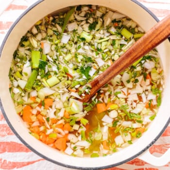 Chopped vegetables floating in water in a soup pot with a wooden spoon alongside.