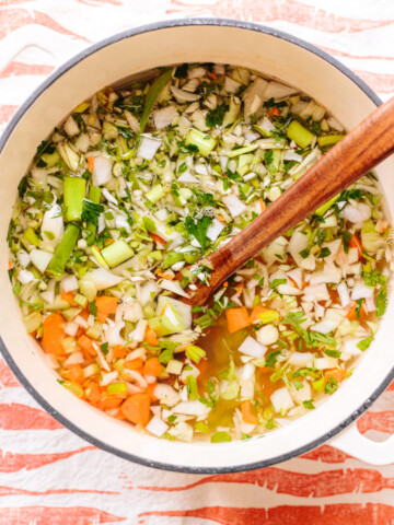 Chopped vegetables floating in water in a soup pot with a wooden spoon alongside.