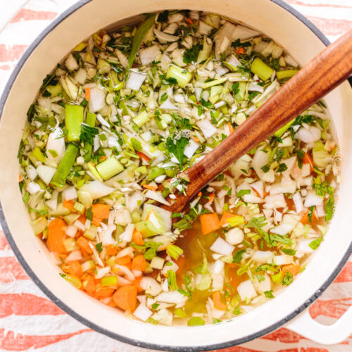 Chopped vegetables floating in water in a soup pot with a wooden spoon alongside.