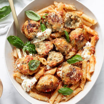 A bowl of penne pasta topped with golden-brown ricotta meatballs, dollops of creamy ricotta cheese, fresh basil leaves, and a sprinkle of grated cheese, with a fork resting on the side.
