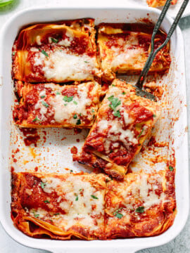 A baking dish filled with sliced cheese lasagna topped with melted cheese and tomato sauce, with a serving spatula lifting one piece. Fresh herbs are sprinkled on top.