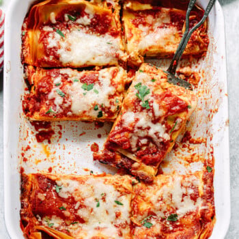 A baking dish filled with sliced cheese lasagna topped with melted cheese and tomato sauce, with a serving spatula lifting one piece. Fresh herbs are sprinkled on top.