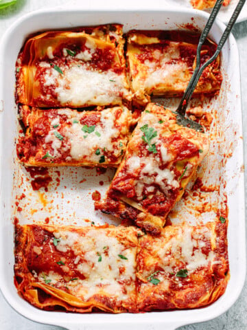 A baking dish filled with sliced cheese lasagna topped with melted cheese and tomato sauce, with a serving spatula lifting one piece. Fresh herbs are sprinkled on top.