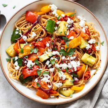 A bowl of veggie pasta topped with roasted vegetables—zucchini, red onion, and bell peppers—garnished with crumbled feta cheese and fresh herbs. A beige napkin and a small plate of feta are nearby.