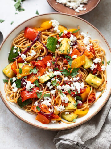 A bowl of veggie pasta topped with roasted vegetables—zucchini, red onion, and bell peppers—garnished with crumbled feta cheese and fresh herbs. A beige napkin and a small plate of feta are nearby.