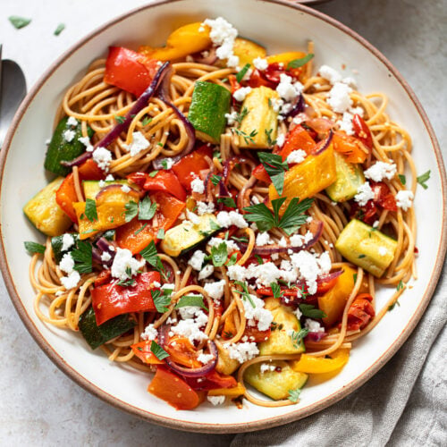 A bowl of veggie pasta topped with roasted vegetables—zucchini, red onion, and bell peppers—garnished with crumbled feta cheese and fresh herbs. A beige napkin and a small plate of feta are nearby.