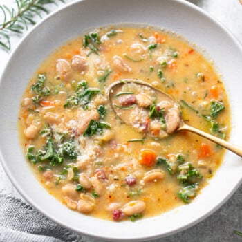 A bowl of creamy Tuscan white bean soup with kale, carrots, and herbs, topped with grated cheese. A gold spoon rests in the bowl. A sprig of rosemary and a small dish of red pepper flakes are nearby.