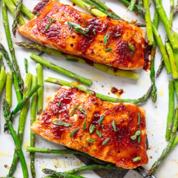Two glazed broiled salmon fillets topped with chopped scallions on a sheet pan with asparagus spears.