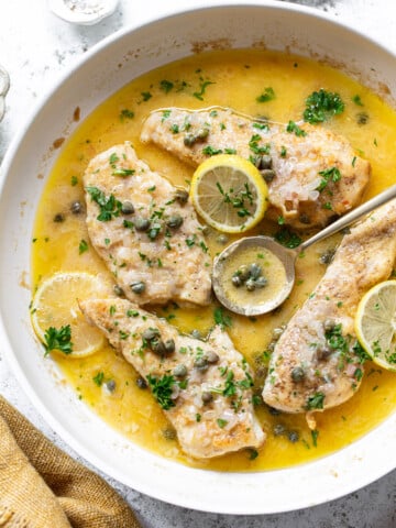 Chicken breasts in a skillet with a lemon-caper sauce, reminiscent of chicken piccata, garnished with parsley and lemon slices. A spoon rests in the sauce, and capers and fresh herbs are sprinkled on top.