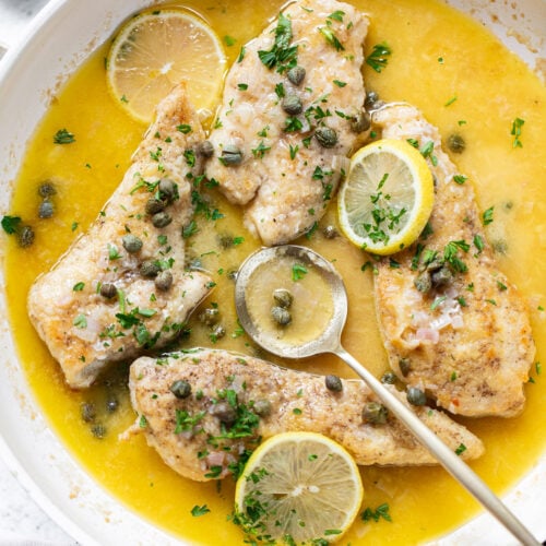 Chicken breasts in a pan surrounded by lemon butter sauce, capers, and chopped parsley.