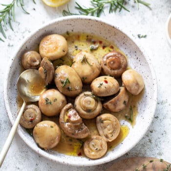 Marinated mushrooms in olive oil and vinegar on a small serving plate, with a spoon on the side, herb sprigs, and lemon wedge in the background.