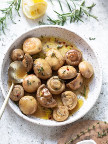 Marinated mushrooms in olive oil and vinegar on a small serving plate, with a spoon on the side, herb sprigs, and lemon wedge in the background.