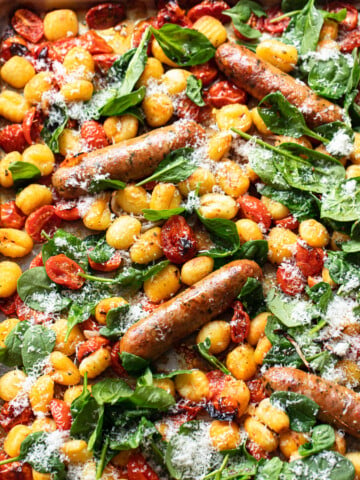 Close-up, overhead photo of roasted Italian sausage and gnocchi sheet pan dinner with grated cheese and spinach leaves.