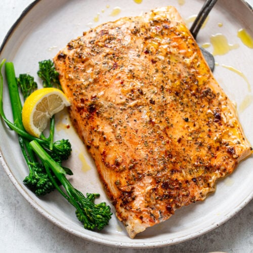 A baked fillet of salmon on a serving plate topped with a lemon mayo sauce and spices, and garnished with green broccolini and wedge of lemon.