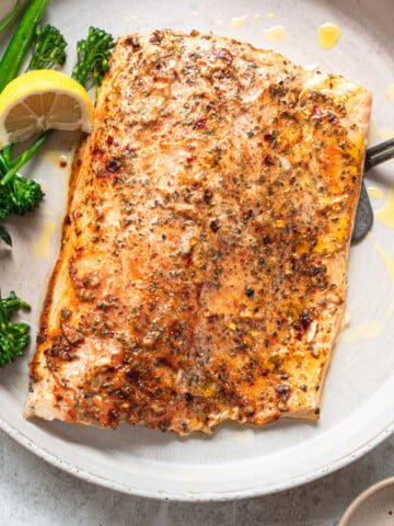 A baked fillet of salmon on a serving plate topped with a lemon mayo sauce and spices, and garnished with green broccolini and wedge of lemon.