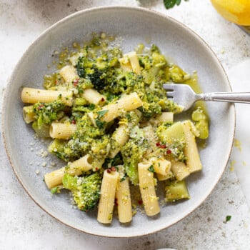 A bowl of broccoli pasta features rigatoni tossed with broccoli, grated cheese, and red pepper flakes, garnished with parsley. A fork rests in the bowl, with lemon, cheese, and fresh herbs nearby.