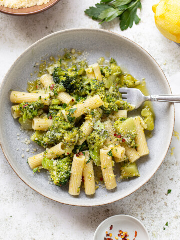 A bowl of broccoli pasta features rigatoni tossed with broccoli, grated cheese, and red pepper flakes, garnished with parsley. A fork rests in the bowl, with lemon, cheese, and fresh herbs nearby.