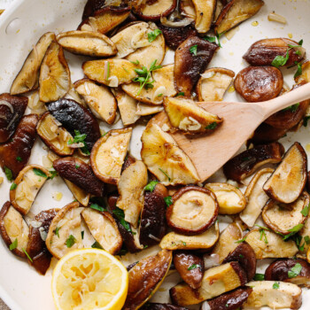 Cooked slices of shiitake mushrooms and a lemon wedge in a saute pan, with a wooden spoon on the side of the pan stirring them.