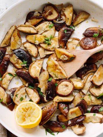 Cooked slices of shiitake mushrooms and a lemon wedge in a saute pan, with a wooden spoon on the side of the pan stirring them.