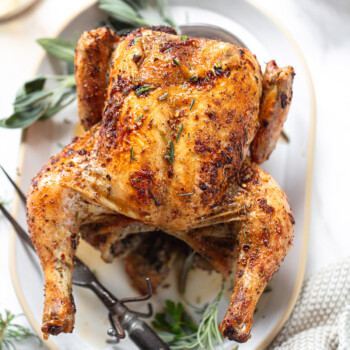 An herb-rubbed roasted beer can chicken on a serving platter, with a serving fork on the side.