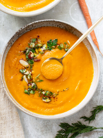 Creamy carrot soup in a serving bowl, with chopped almonds and parsley garnished on top and a spoon.