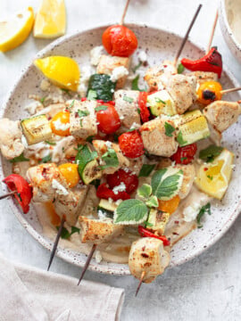 Grilled chicken kabobs and vegetable skewers with tomatoes, zucchini, and peppers are served on a plate with lemon wedges, fresh herbs, and crumbled cheese on a light background.