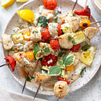 Grilled chicken kabobs and vegetable skewers with tomatoes, zucchini, and peppers are served on a plate with lemon wedges, fresh herbs, and crumbled cheese on a light background.