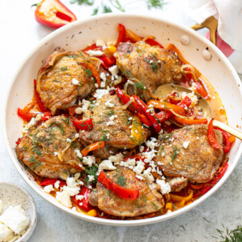 Golden-skinned chicken thighs with red bell pepper sauce, feta cheese, and fresh dill, served in a frying pan.