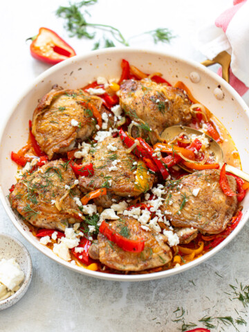 Golden-skinned chicken thighs with red bell pepper sauce, feta cheese, and fresh dill, served in a frying pan.
