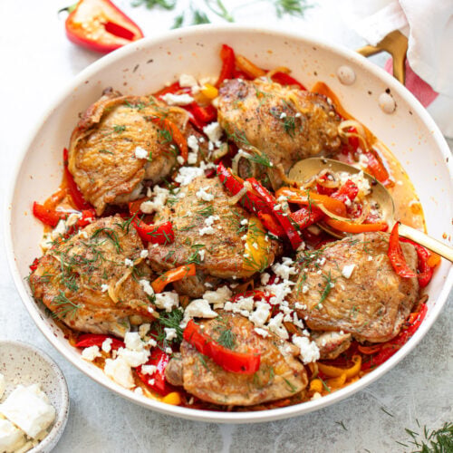 Golden-skinned chicken thighs with red bell pepper sauce, feta cheese, and fresh dill, served in a frying pan.