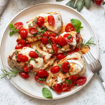 Four servings of chicken breast and melted mozzarella cheese bruschetta on a serving platter, garnished with sprigs of basil and rosemary.