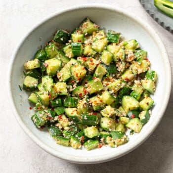 A white bowl filled with Indian cucumber salad, garnished with peanuts minced garlic, red pepper flakes, and fresh herbs on a light gray surface.