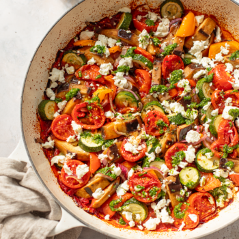 A close-up of a pan filled with colorful baked ratatouille—roasted tomatoes, zucchini, eggplant, and bell peppers—topped with crumbled cheese and green herb sauce on a light background.